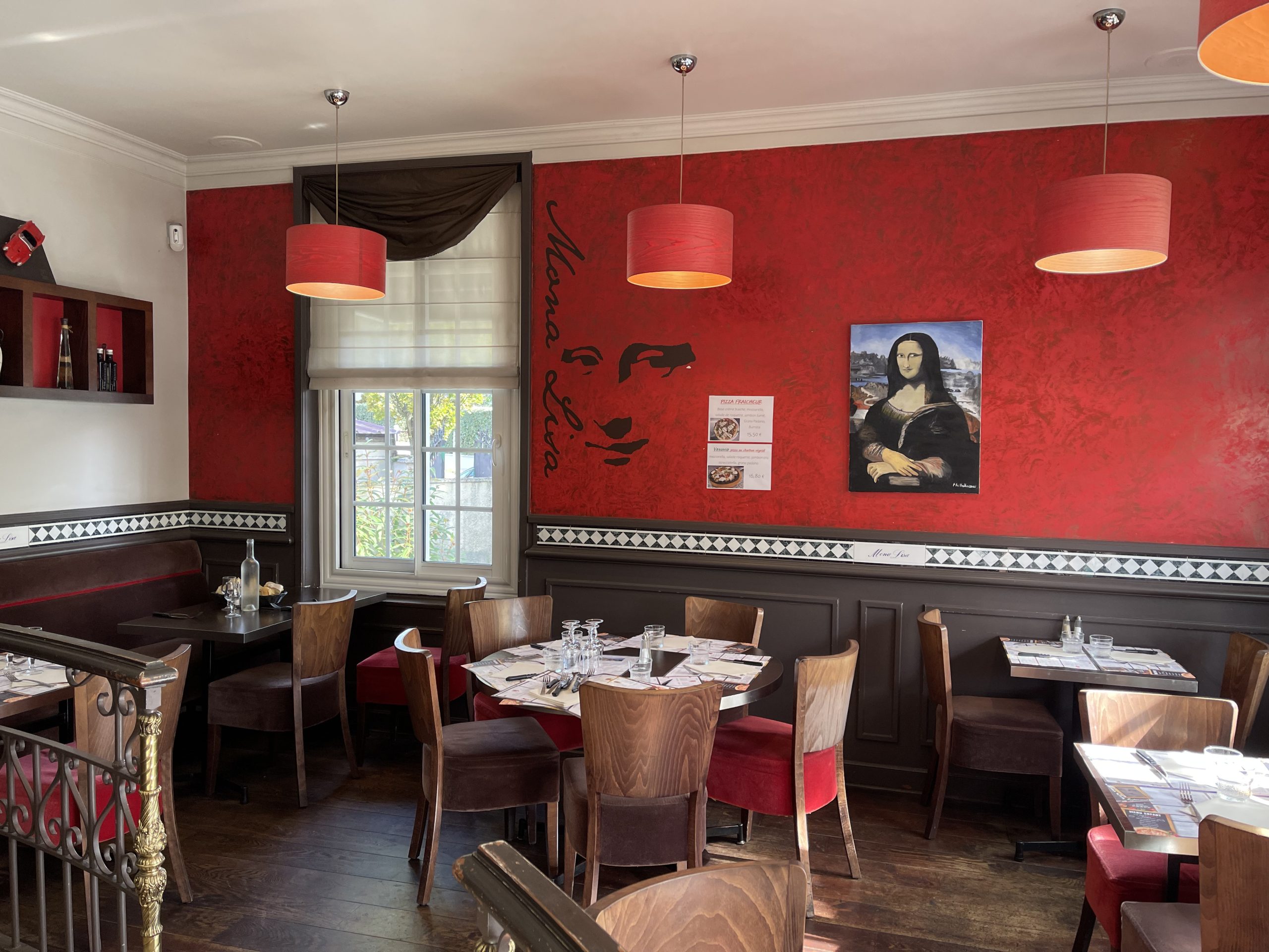 À l'entrée du restaurant Mona Lisa, l'intérieur accueille les visiteurs avec des tables et des chaises en bois. Le mur du fond, peint en rouge, arbore le visage iconique de la Joconde, ajoutant une touche artistique distinctive. Sur le côté droit, une reproduction fidèle du portrait de la Joconde complète ce décor inspiré de l'art italien.