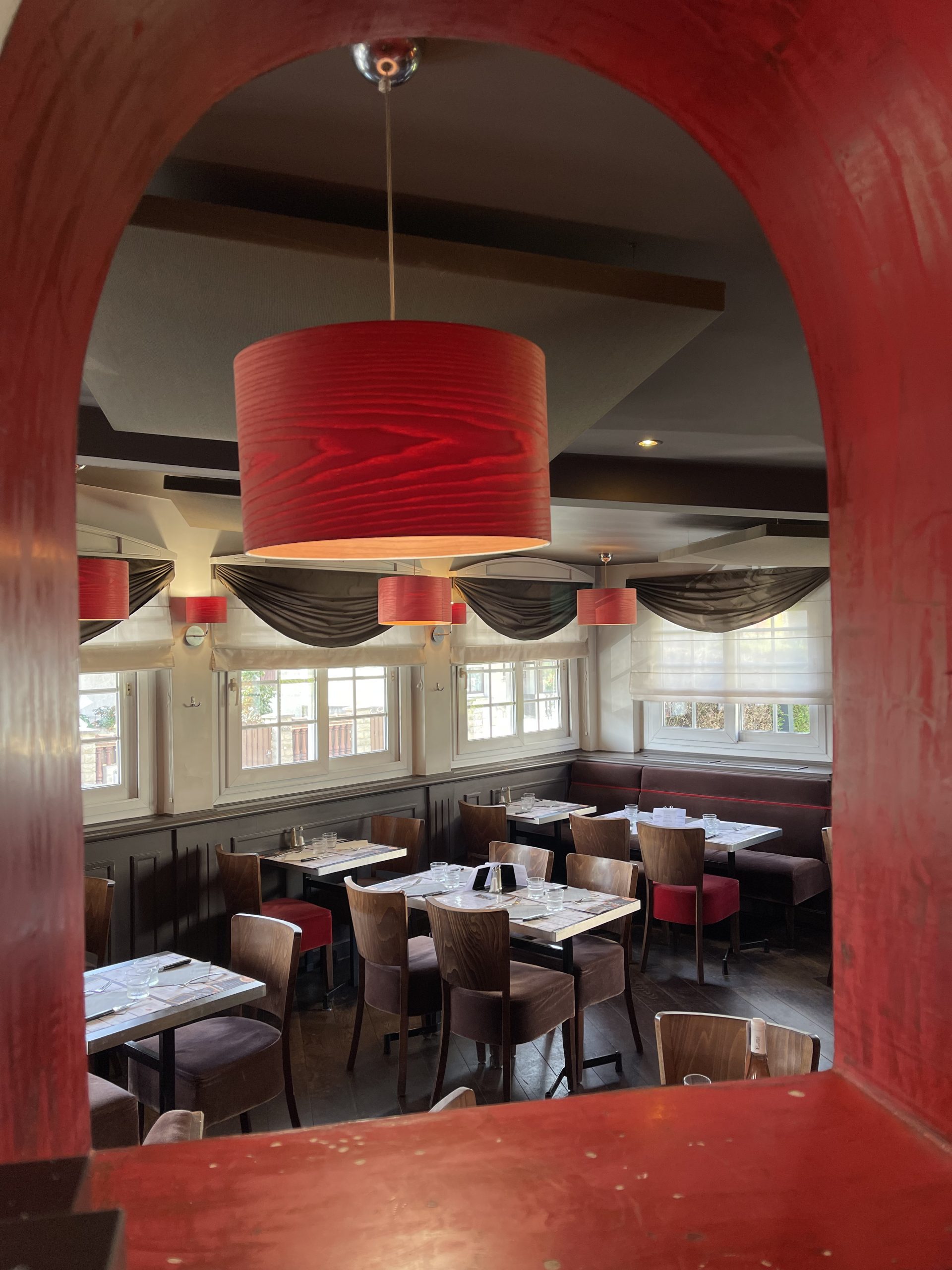 Vue à travers une ouverture en arc dans un mur rouge, révélant l'intérieur chaleureux de la salle principale du restaurant Mona Lisa à Villiers-sur-Marne (94). Les tables et chaises en bois disposées de façon accueillante invitent à la détente et à la dégustation d'un repas italien traditionnel dans un cadre élégant et plein de charme.